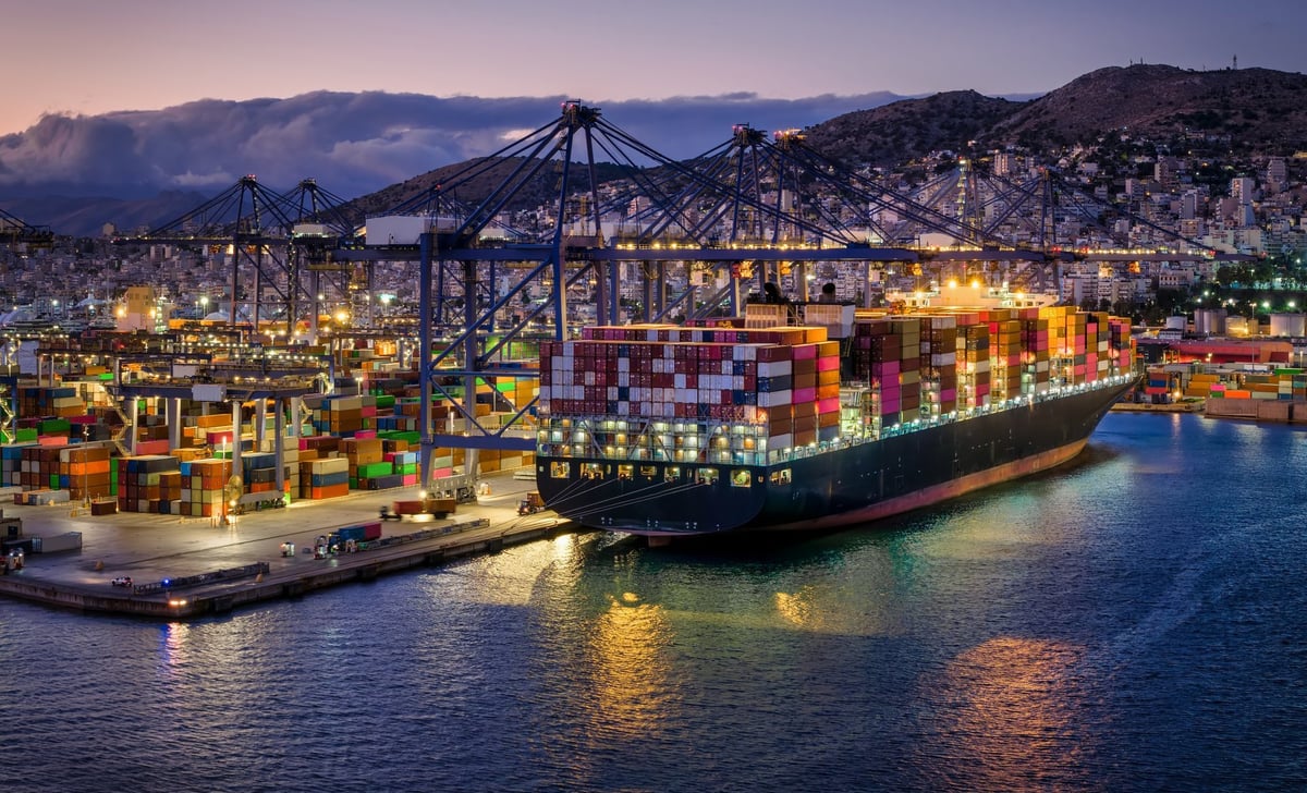 A illuminated, large container super cargo ship being unloaded by cranes and trucks in a commercial terminal during evening